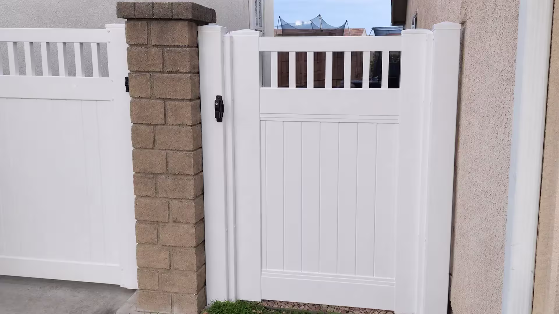 Through the vertical picket top, a distant rooftop shows past this White vinyl single gate set in a stucco passage