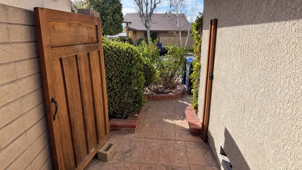 Before installation, an open wood gate leads through a paved passage between a block wall and stucco house toward trimmed hedges.