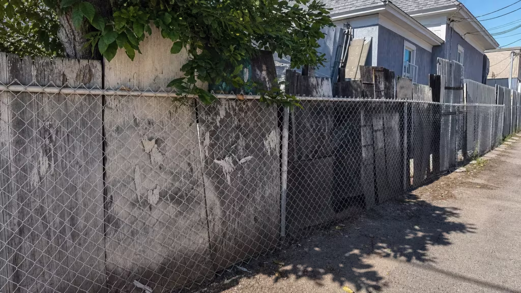 Before installation, chain-link fencing and weathered plywood panels line an alley behind several homes.