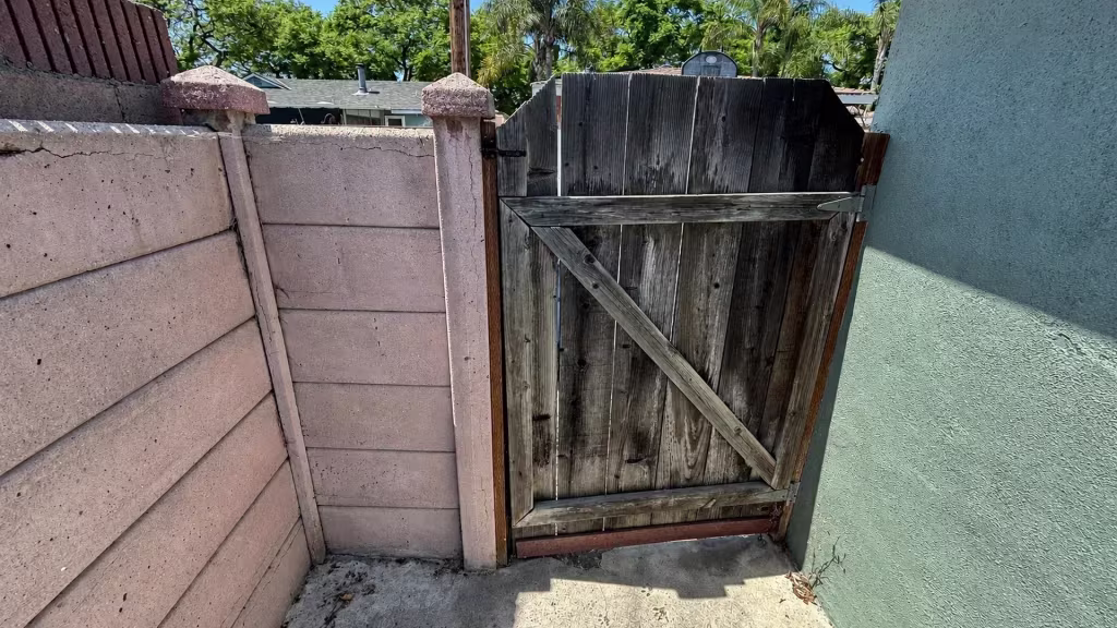 Before installation, a weathered wood gate stands between a pink concrete wall and teal stucco house.
