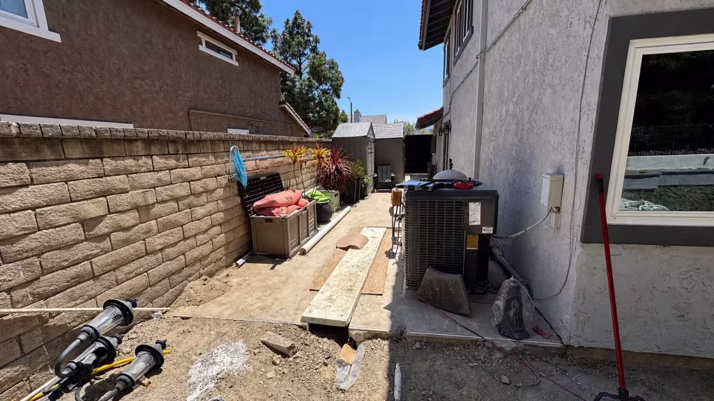 Before installation, an unfinished side yard contains an air-conditioning unit, storage sheds, boards, and construction equipment beside a tan block wall.