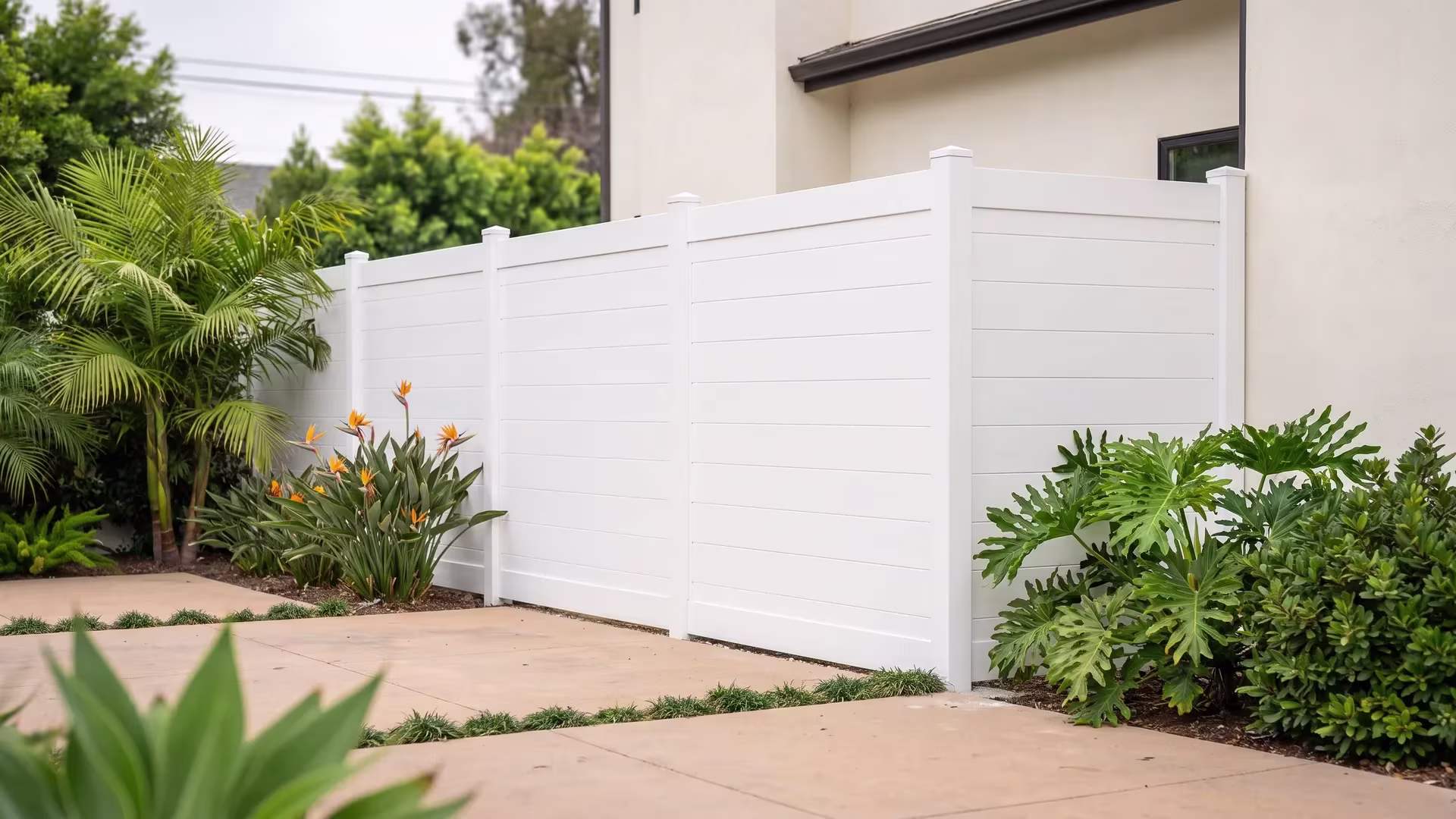 White aluminum privacy fence meeting a building wall beside warm concrete pavers and dense shrubs