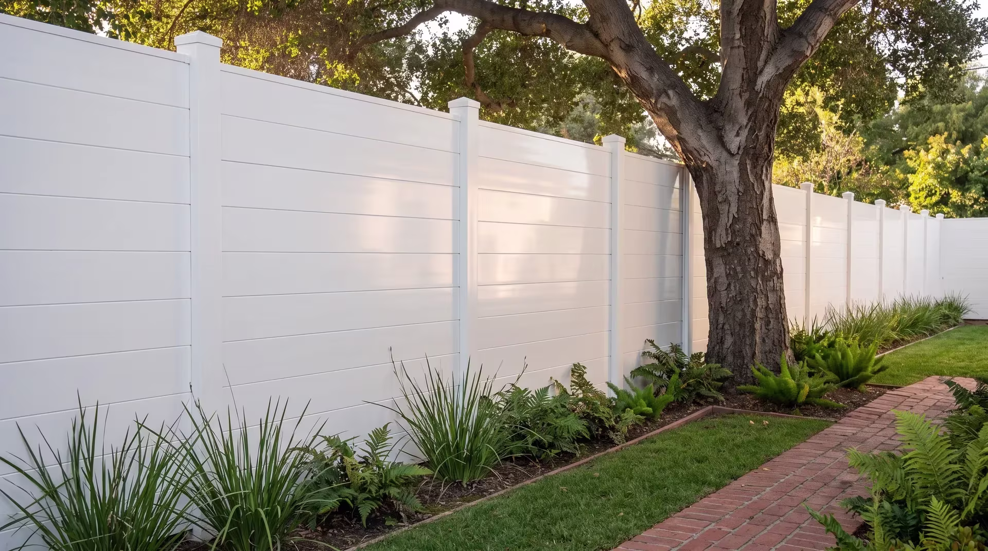 A mature shade tree anchors this White aluminum privacy fence running level past a brick-edged garden path