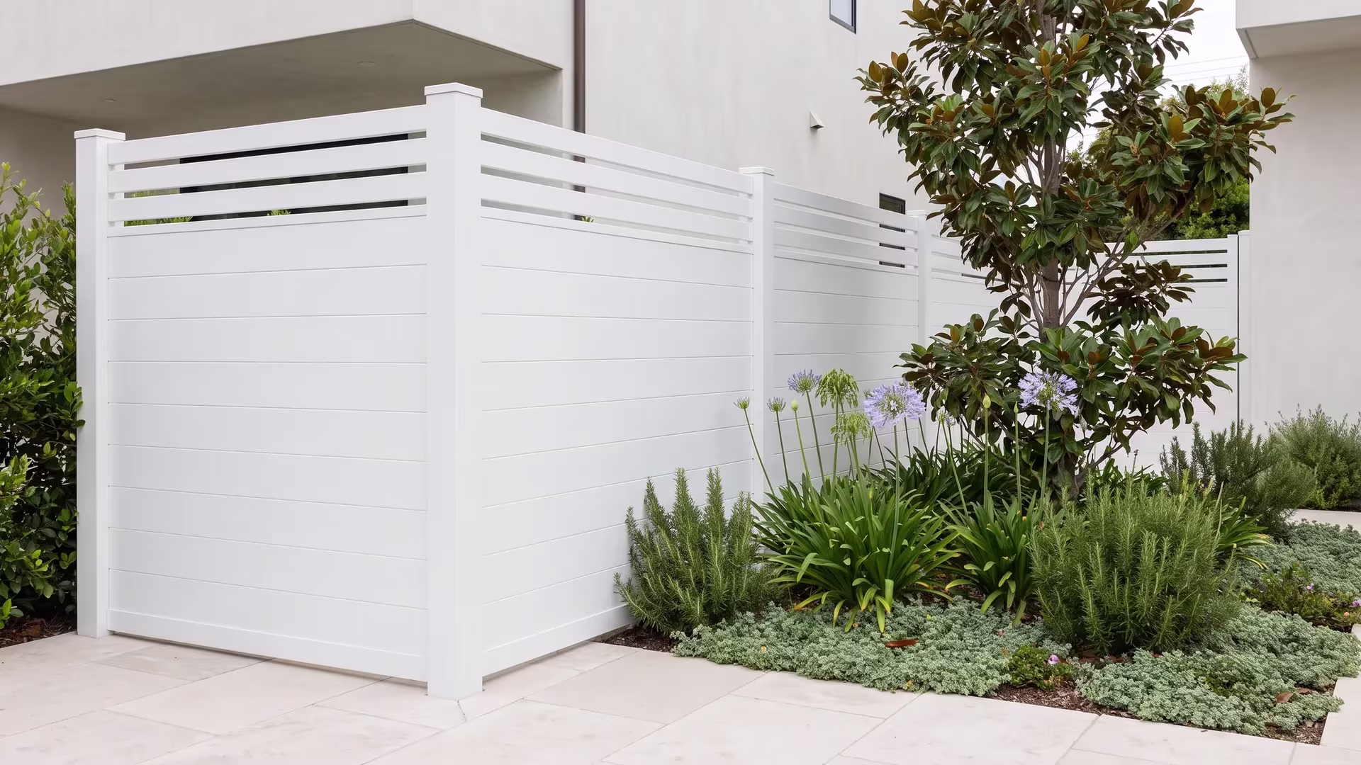 This White aluminum privacy fence borders a courtyard's pale stone paving near magnolia
