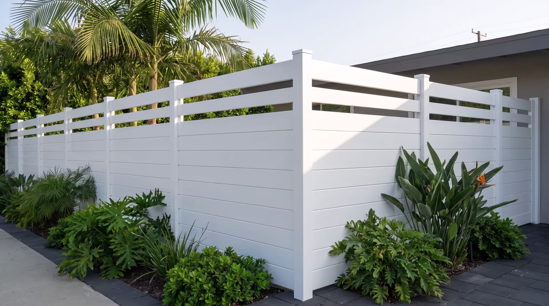 A ranch home's low roofline overlooks this White aluminum privacy fence turning near dense green shrubs