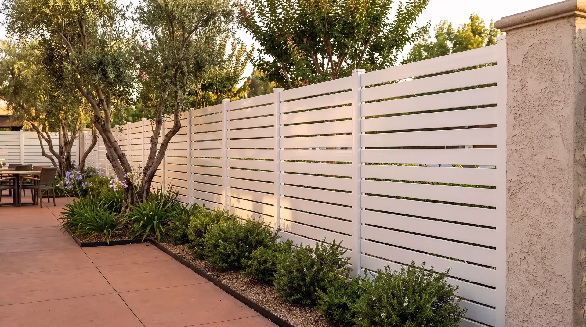 Olive trees shade a paved terrace where a White aluminum semi privacy fence recedes toward a masonry column