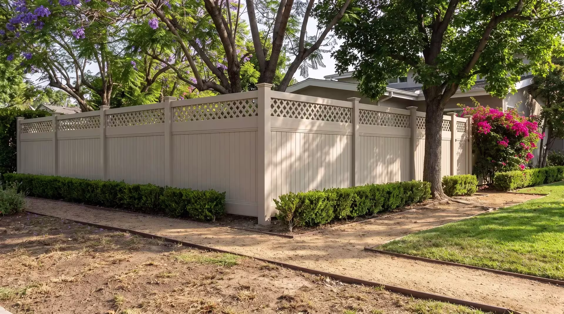 Tan vinyl privacy fence crowned by a lattice course runs beside steel-edged granite paths under a jacaranda canopy