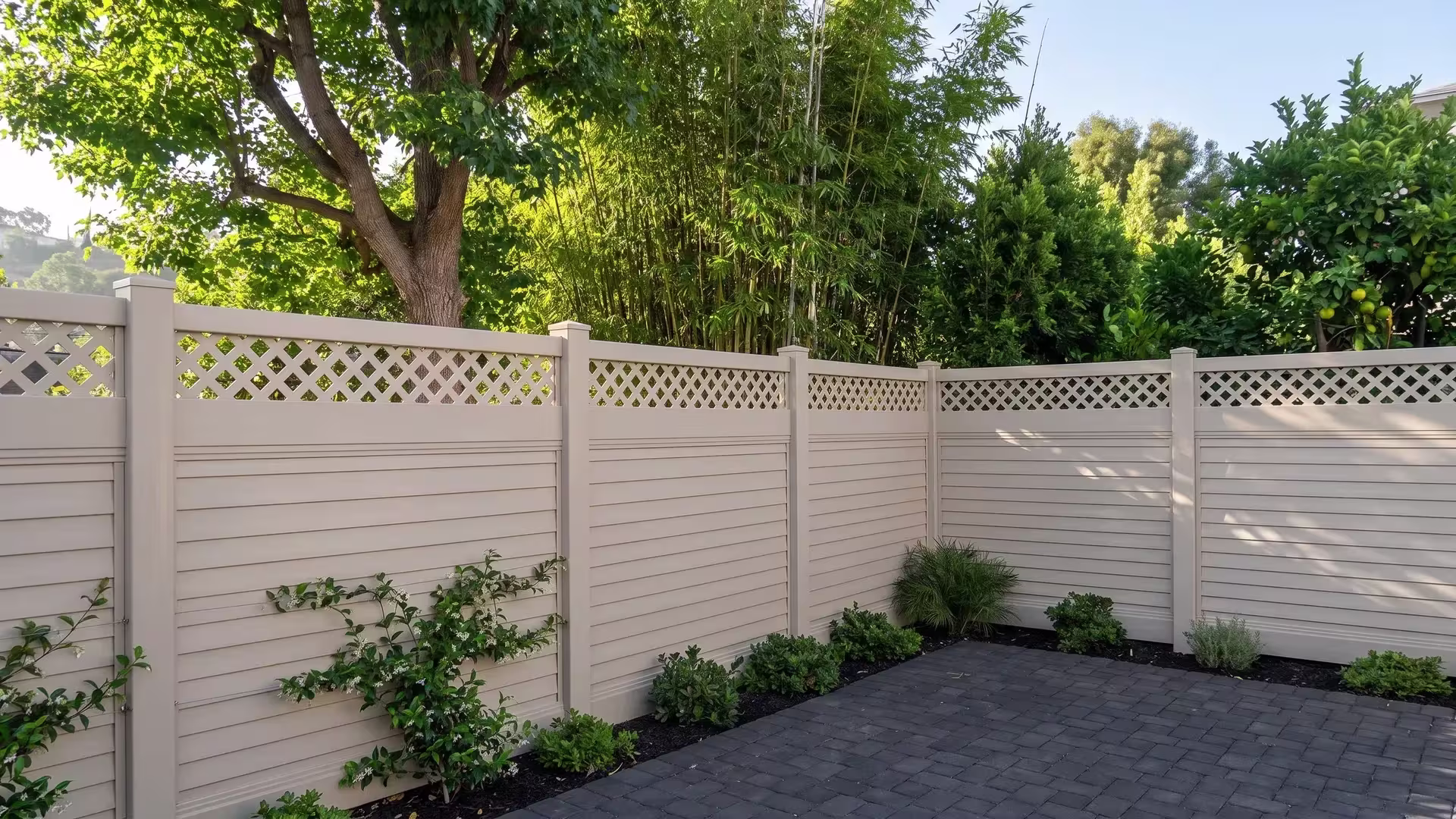 Tan vinyl horizontal privacy fence with a lattice top set along a hillside backyard with dark charcoal pavers