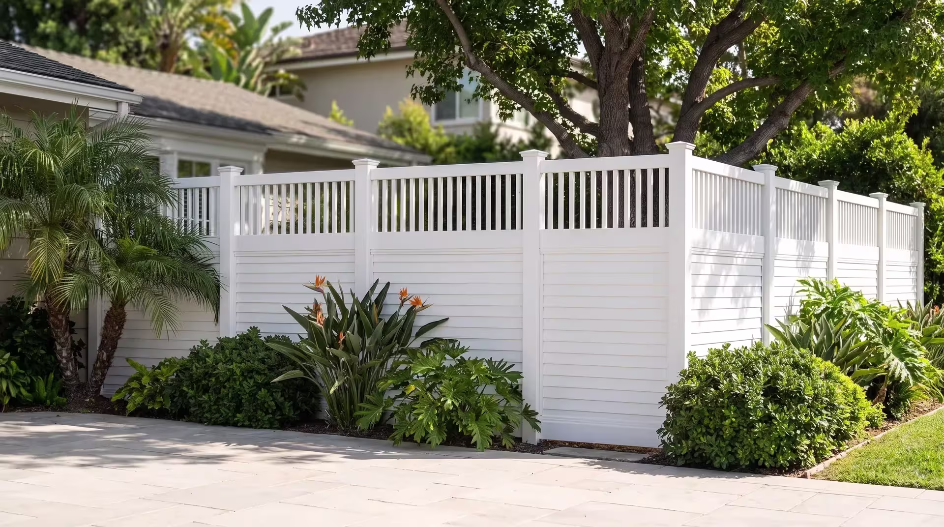 Pale stone paving beneath queen palms borders a White vinyl fence, horizontal panels finished with picket top trim