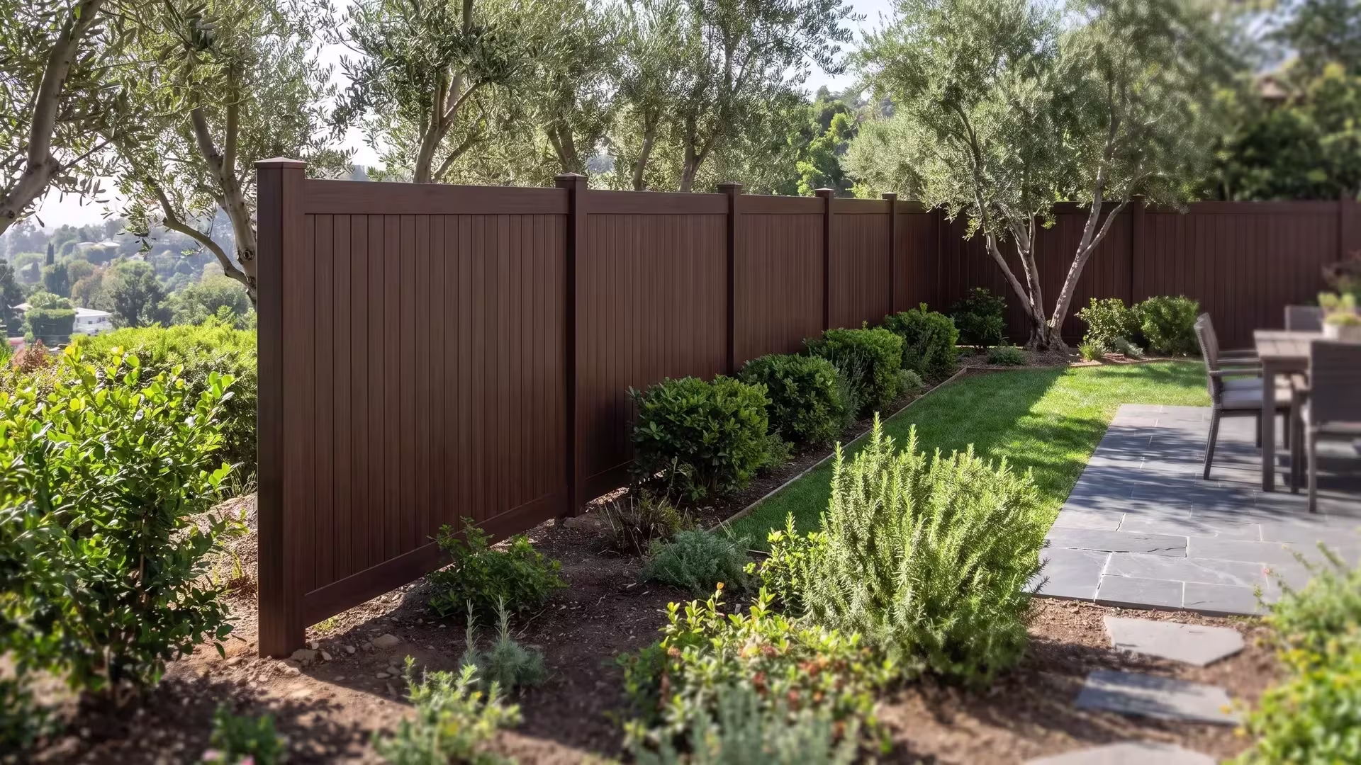 Dark Sequoia vinyl vertical privacy fence borders an outdoor dining terrace beside olive trees and rosemary